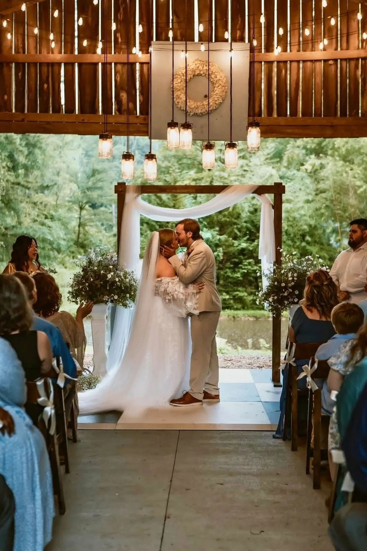 Bride and groom kissing beneath a draped wooden arbor as their guests look on